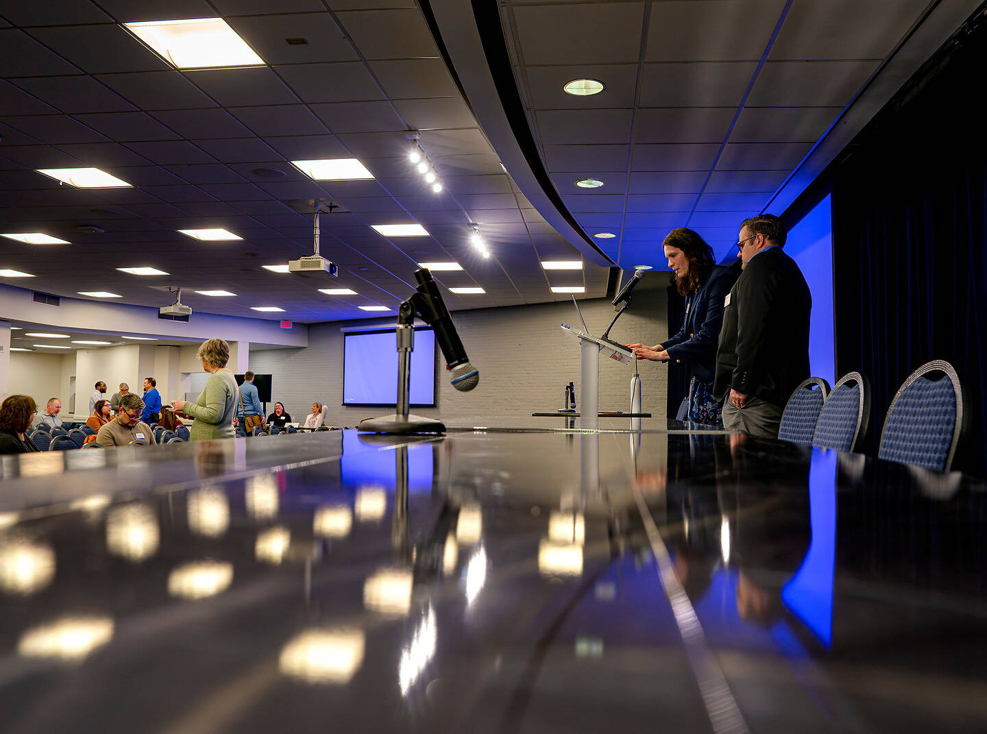 Two individuals stand at a podium in a conference room, addressing an audience seated around a long reflective table.